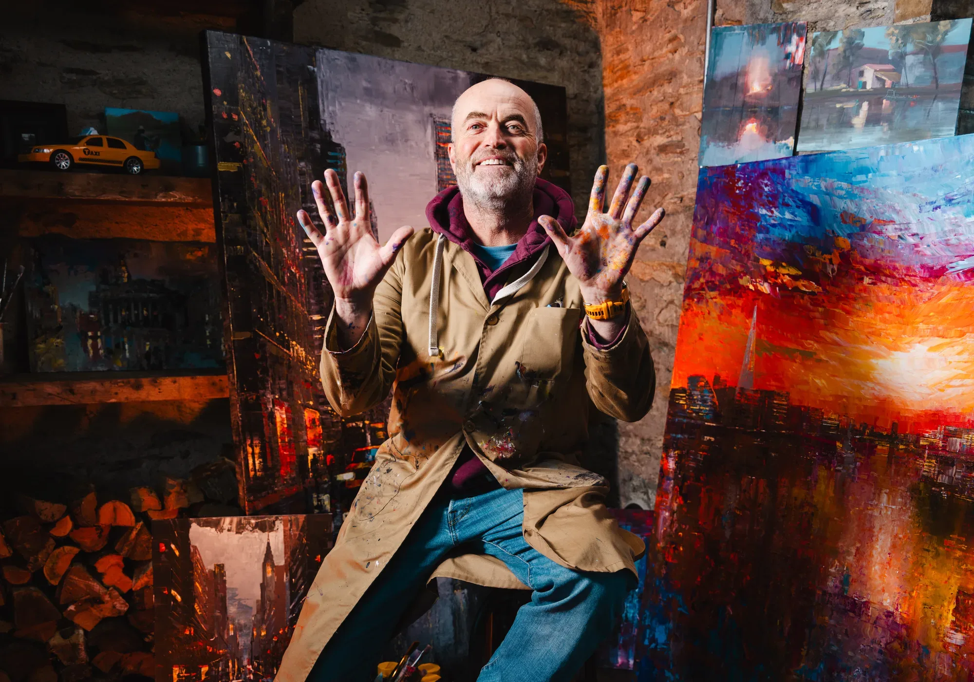 Paul Kenton in his studio, smiling and holding up his paint-covered hands among his paintings